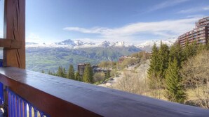 Un balcón con vistas a montañas nevadas, una zona boscosa y un edificio de varios pisos.