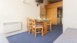 Un petit coin repas avec une table et des chaises, un téléviseur fixé au mur et un espace cuisine visible à travers une porte ouverte.