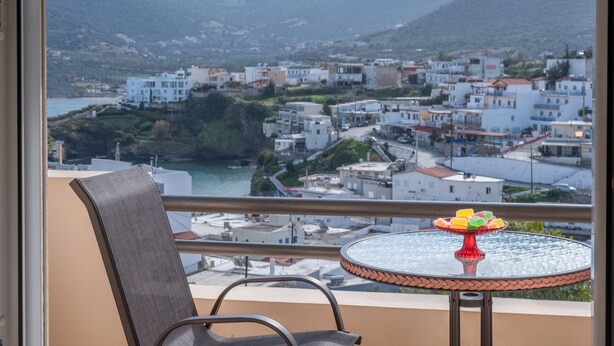 Un balcón con una silla y una mesa con una bandeja de frutas, con vistas a una ciudad costera y montañas al fondo.