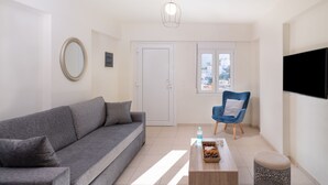 Una sala moderna con un sofá gris, un sillón azul, una mesita de centro con aperitivos, un televisor montado en la pared y una puerta que da a un balcón.