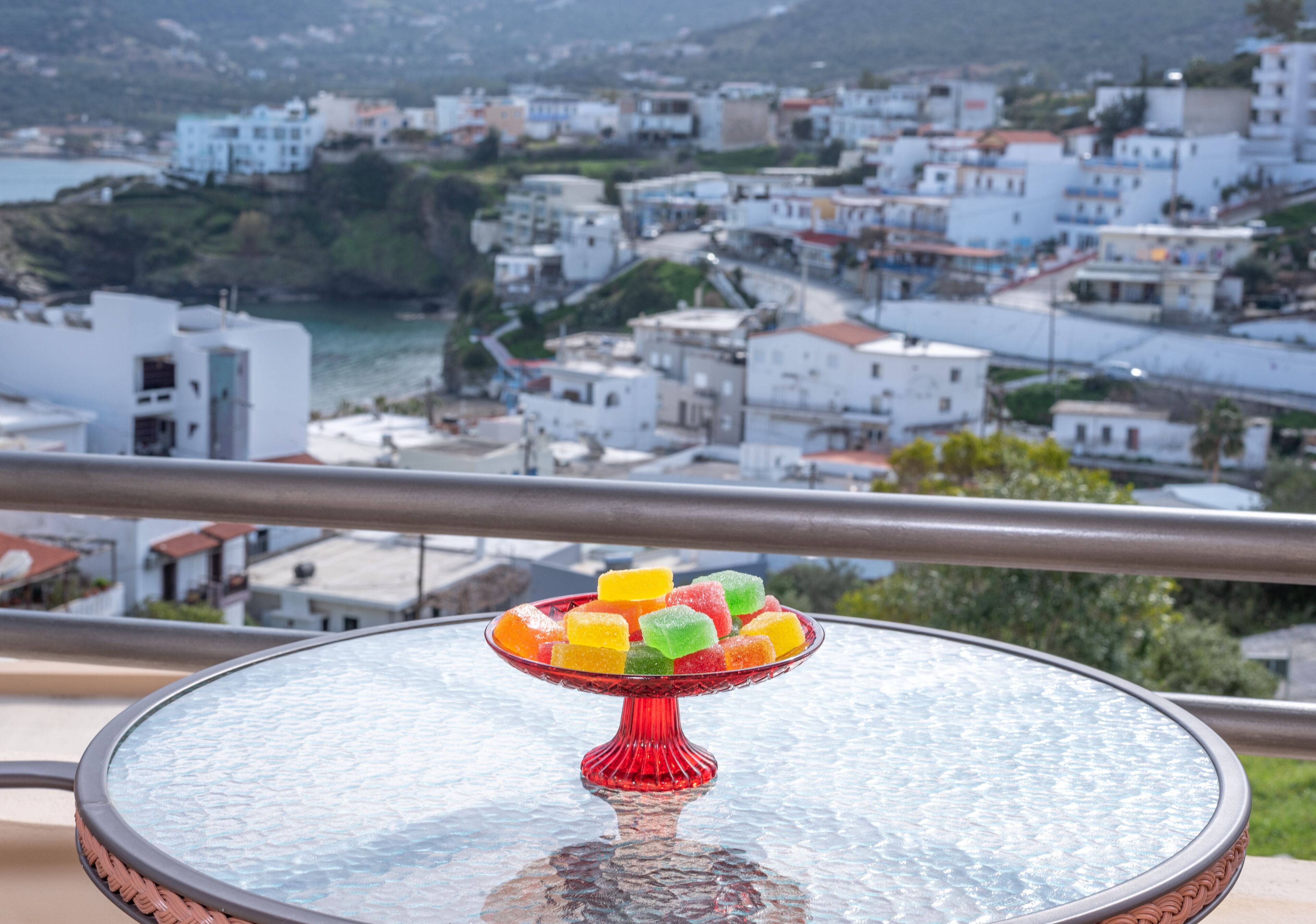 A table with a glass top and a red stand, placed on a balcony overlooking a coastal town with white buildings and a view of the sea.