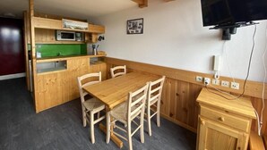 A compact kitchen with wooden cabinets, a dining table, and chairs. A television is mounted on the wall.