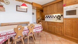 Een gezellige keuken met houten kastjes, een eettafel met stoelen, een rode hanglamp en een klok aan de muur.