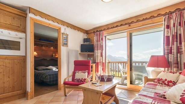 Een gezellige kamer met een houten plafond, een bed met een geruite dekbedovertrek, een rode stoel, een houten tafel en een balkon met uitzicht op de bergen.