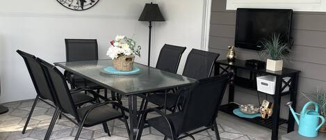 Um alpendre coberto com mesa e cadeiras, uma televisão fixada na parede e uma peça de arte decorativa com o tema do sol.