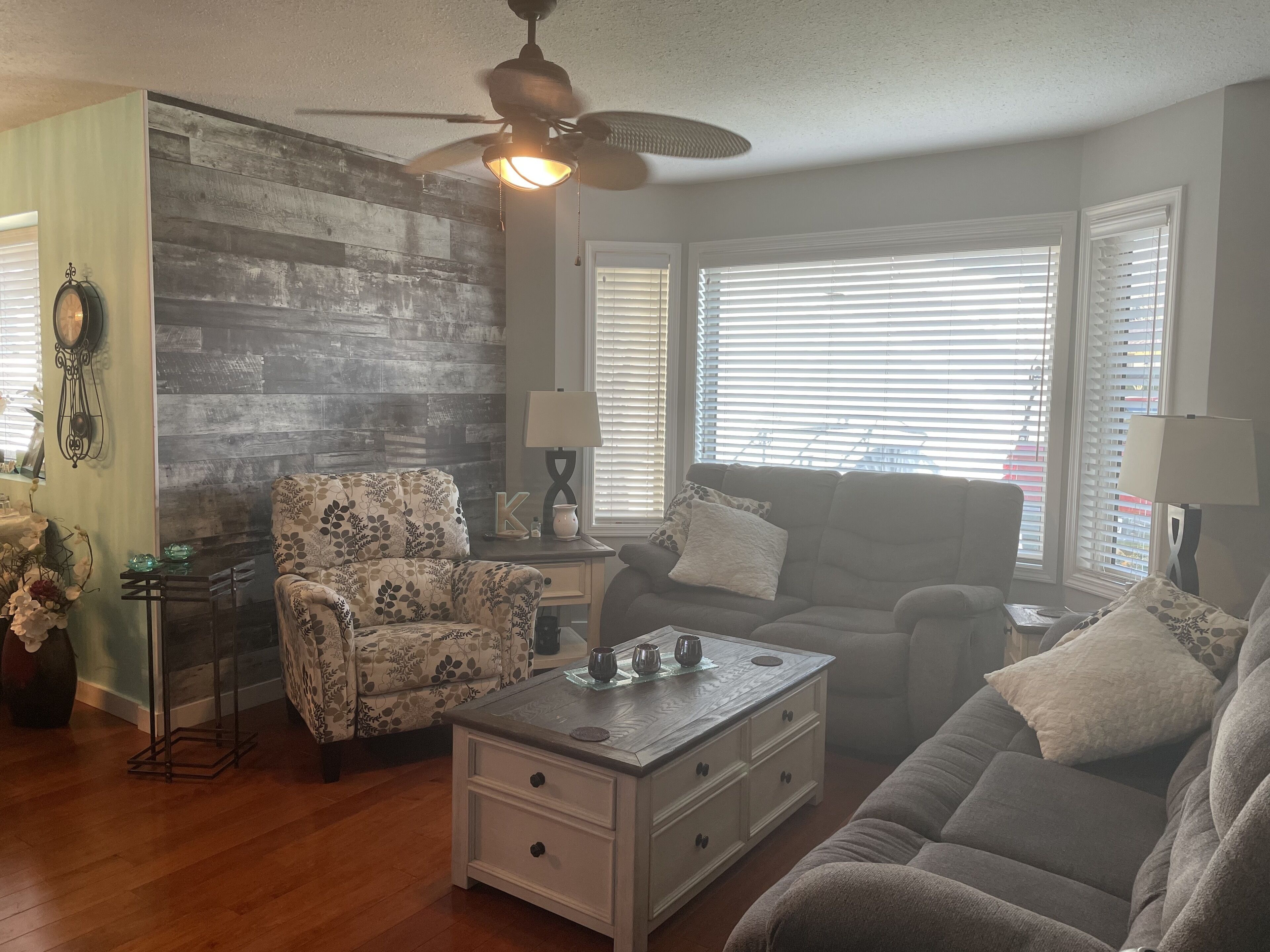 A living room with a wooden accent wall, a ceiling fan, a floral-patterned armchair, a gray sofa, and a coffee table.