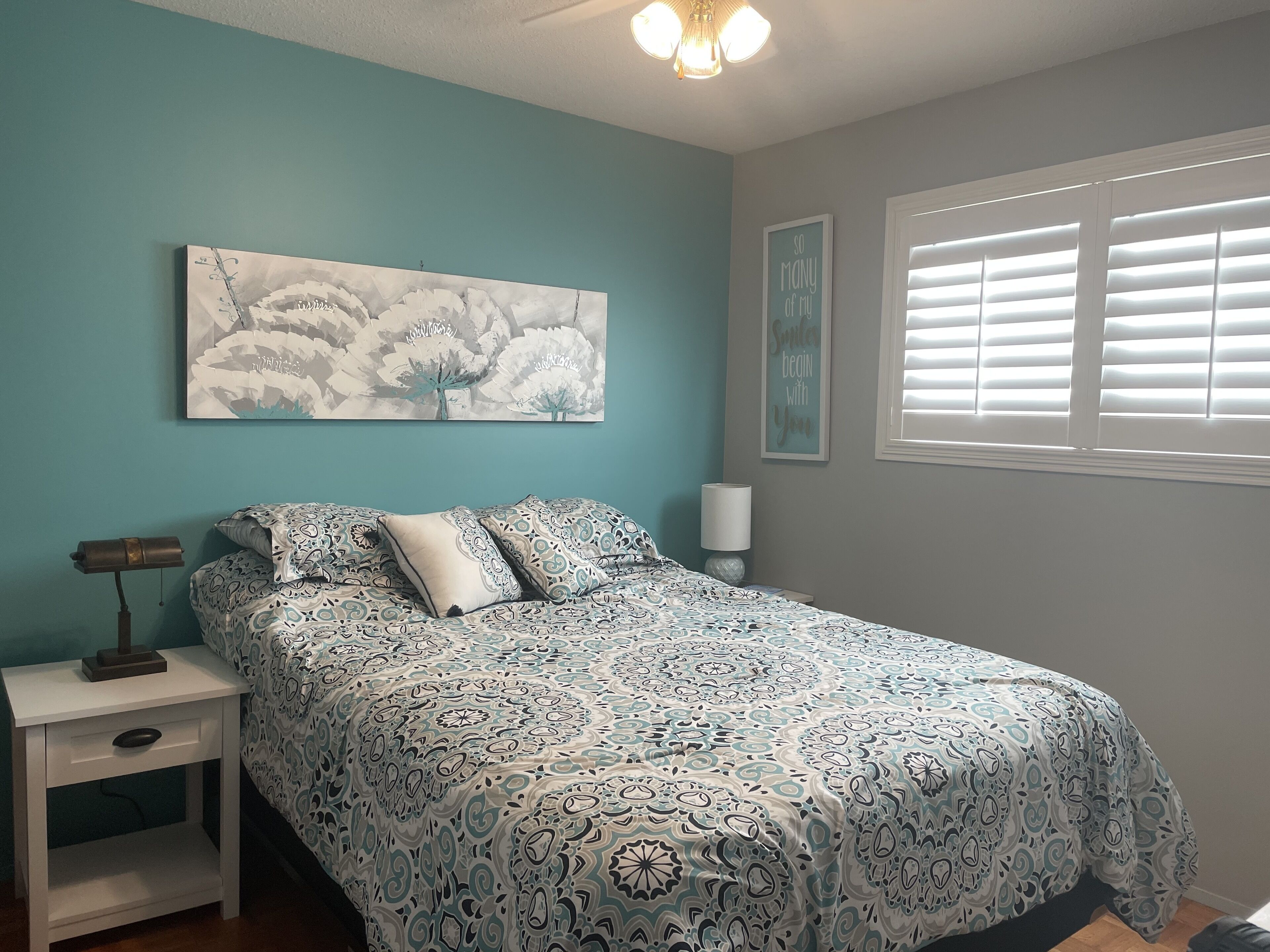 A bedroom with a bed, a nightstand, a lamp, a wall art piece, and a window with blinds.