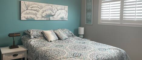 A bedroom with a bed, a nightstand, a lamp, a wall art piece, and a window with blinds.