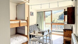 A compact room with bunk beds, a dining table with chairs, a small kitchen area, and a large window offering a view of the outdoors. - 100m from ski slopes, 12nd floor, view ski slopes, swimming pool, balcony, tv, ski locker, 22m² (Le Corbier)