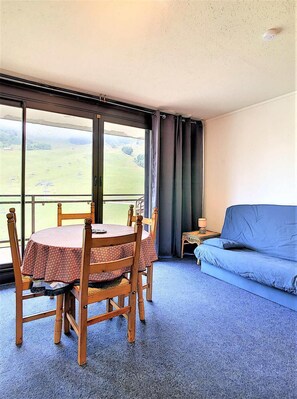 A room with a round wooden table and chairs, a blue sofa, and a large window with a view of a green landscape. - 10m from resort center, 10th floor, view ski slopes, swimming pool, balcony, tv, ski locker, 31m² (Le Corbier)