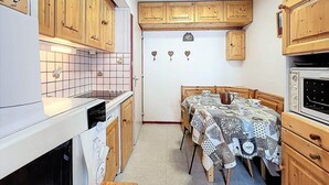 A compact kitchen with wooden cabinets, a dining table with a patterned tablecloth, and a built-in oven. - 10m from ski slopes, 6th floor, view mountain, swimming pool, tv, ski locker, 26m², Le Corbier (Le Corbier)