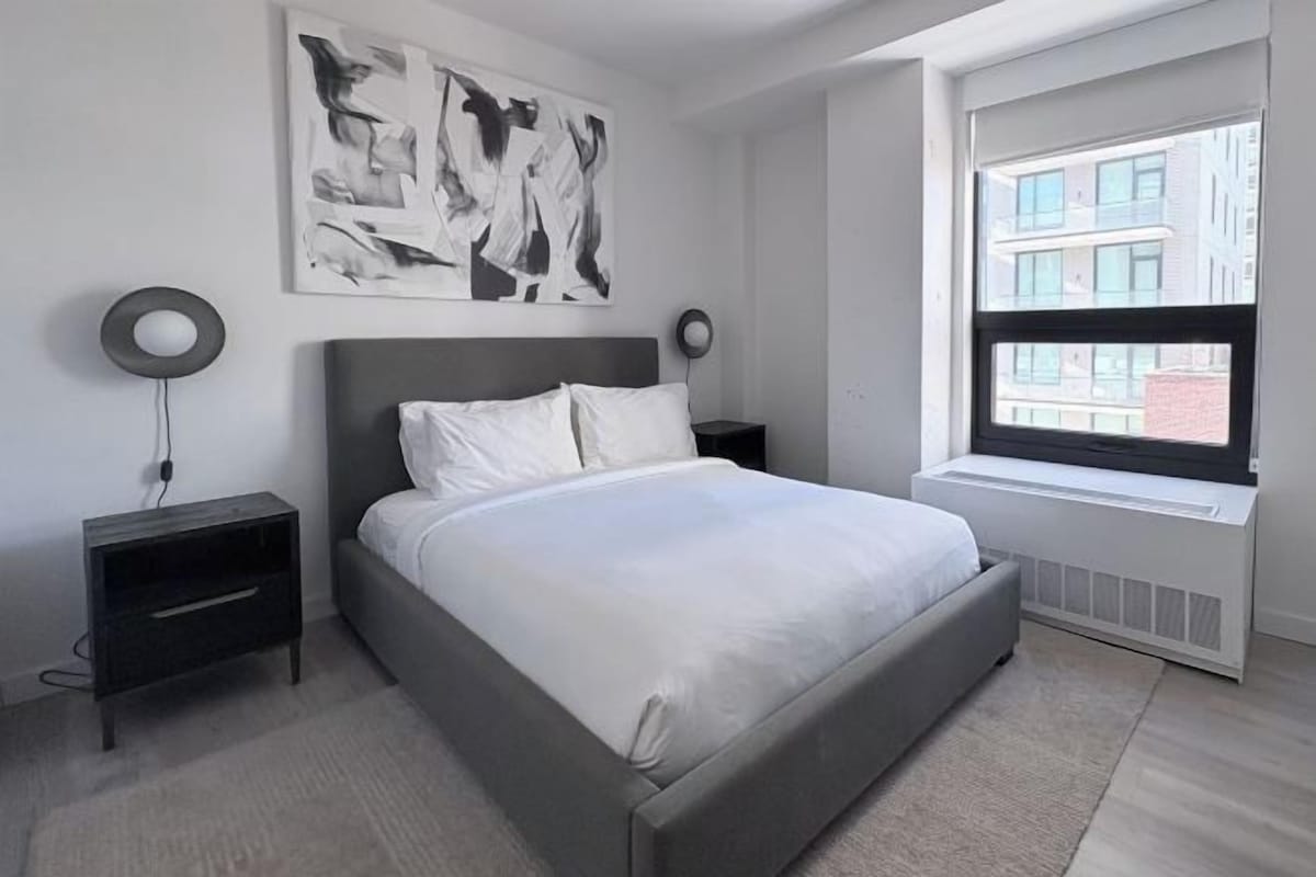 A modern bedroom with a grey bed, white bedding, a large abstract painting, two bedside lamps, a window with a view of buildings, and a radiator.