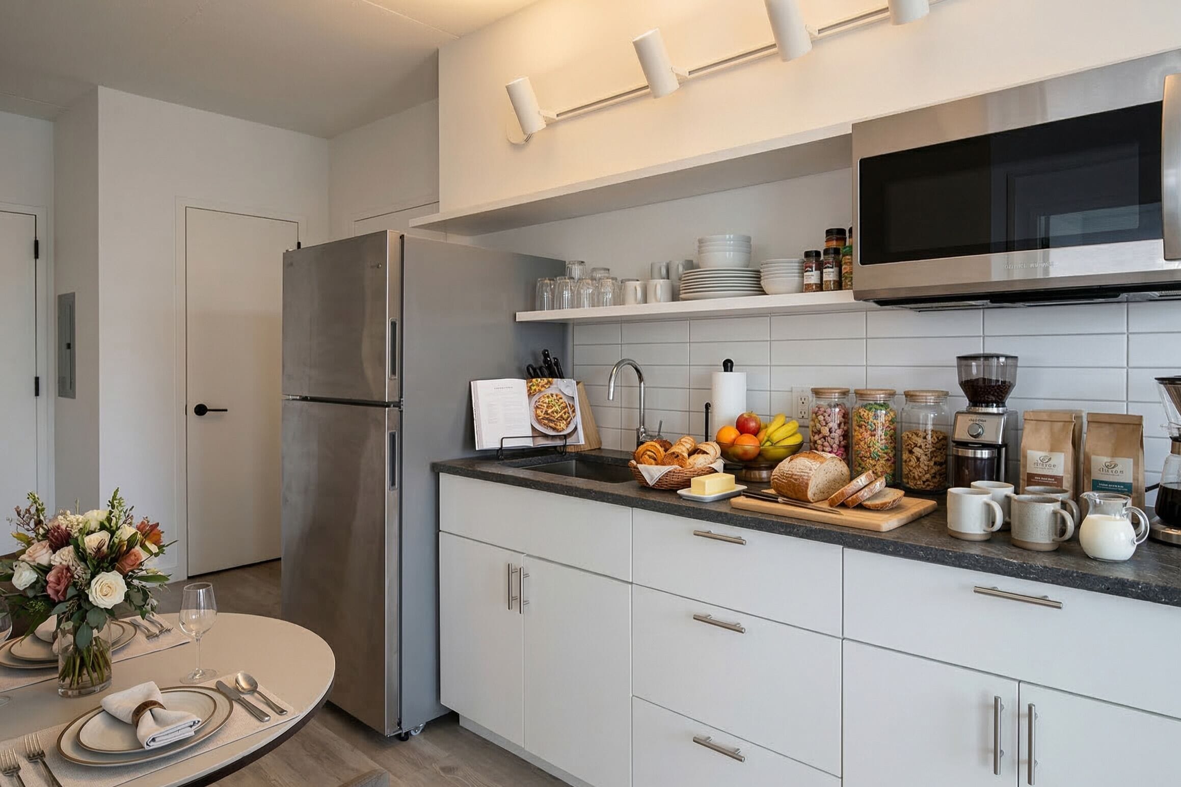Una cucina moderna con frigorifero in acciaio inossidabile, forno a microonde e pensili bianchi. Sul piano di lavoro si trovano diversi alimenti e utensili da cucina.