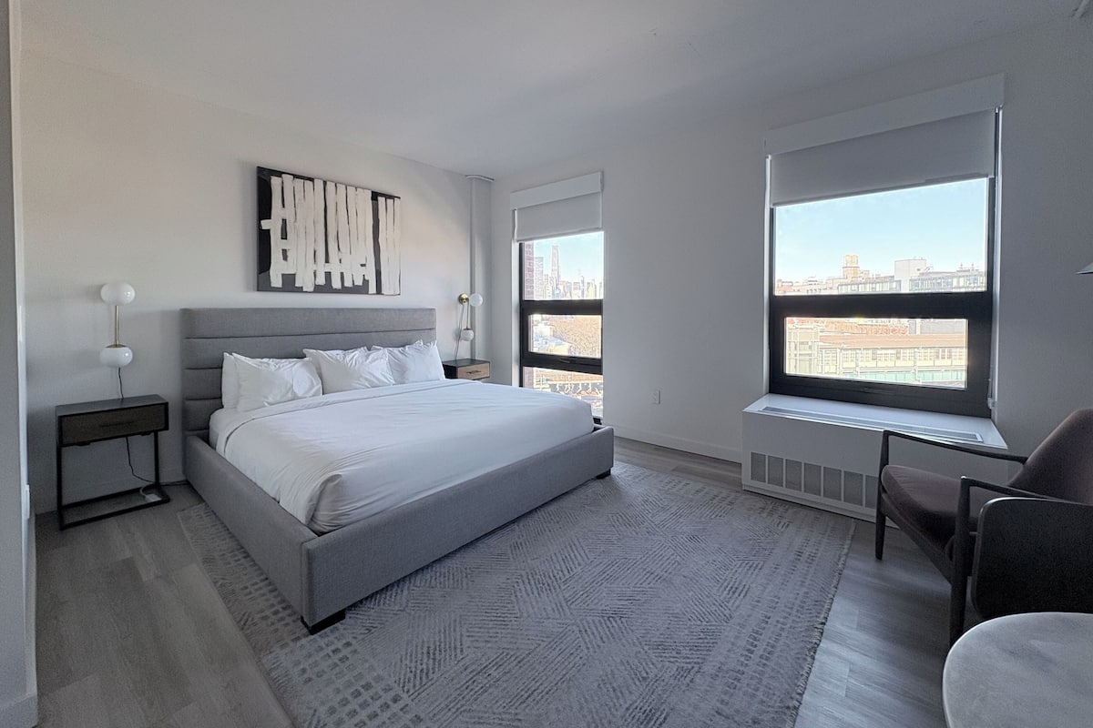 Ein modernes Schlafzimmer mit einem großen Bett, einem Nachttisch, einem Sessel und einem Fenster mit Blick auf die Stadt.