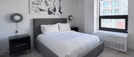 A modern bedroom with a large bed, a wall-mounted abstract painting, two bedside tables with lamps, and a window with a city view.