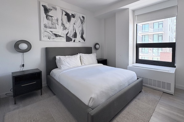 A modern bedroom with a large bed, a wall-mounted abstract painting, two bedside tables with lamps, and a window with a city view.