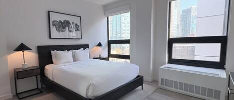 A modern bedroom with a large bed, two bedside tables with lamps, a framed artwork on the wall, and a window with a view of buildings.