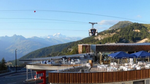 Un restaurant doté d’une terrasse offrant une vue sur une chaîne de montagnes, un téléphérique et une route avec un panneau de signalisation rouge.