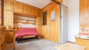 Une pièce de taille modeste aux murs en bois, avec un lit recouvert d’une couverture rouge, une petite table et une fenêtre ornée de rideaux.