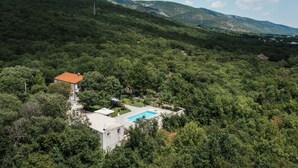 Une maison avec une piscine, entourée d’une forêt dense, située sur une colline avec des montagnes en arrière-plan.