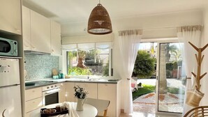 Een moderne keuken met witte kasten, een eethoek met een ronde tafel en stoelen, en uitzicht op een tuin door grote ramen.