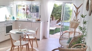 Een moderne keuken met witte kastjes, een eethoek met rieten stoelen en uitzicht op een tuin door grote ramen.