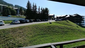 Une vue depuis un balcon donnant sur un parking où se trouvent des voitures, une colline verdoyante et des bâtiments en arrière-plan.