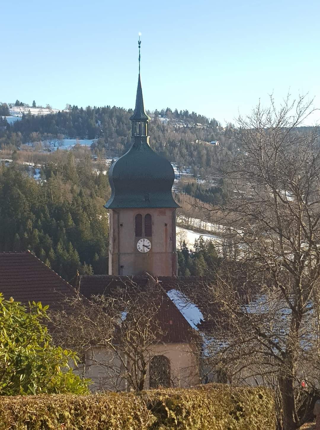A church with a clock tower, surrounded by trees and a hilly landscape.