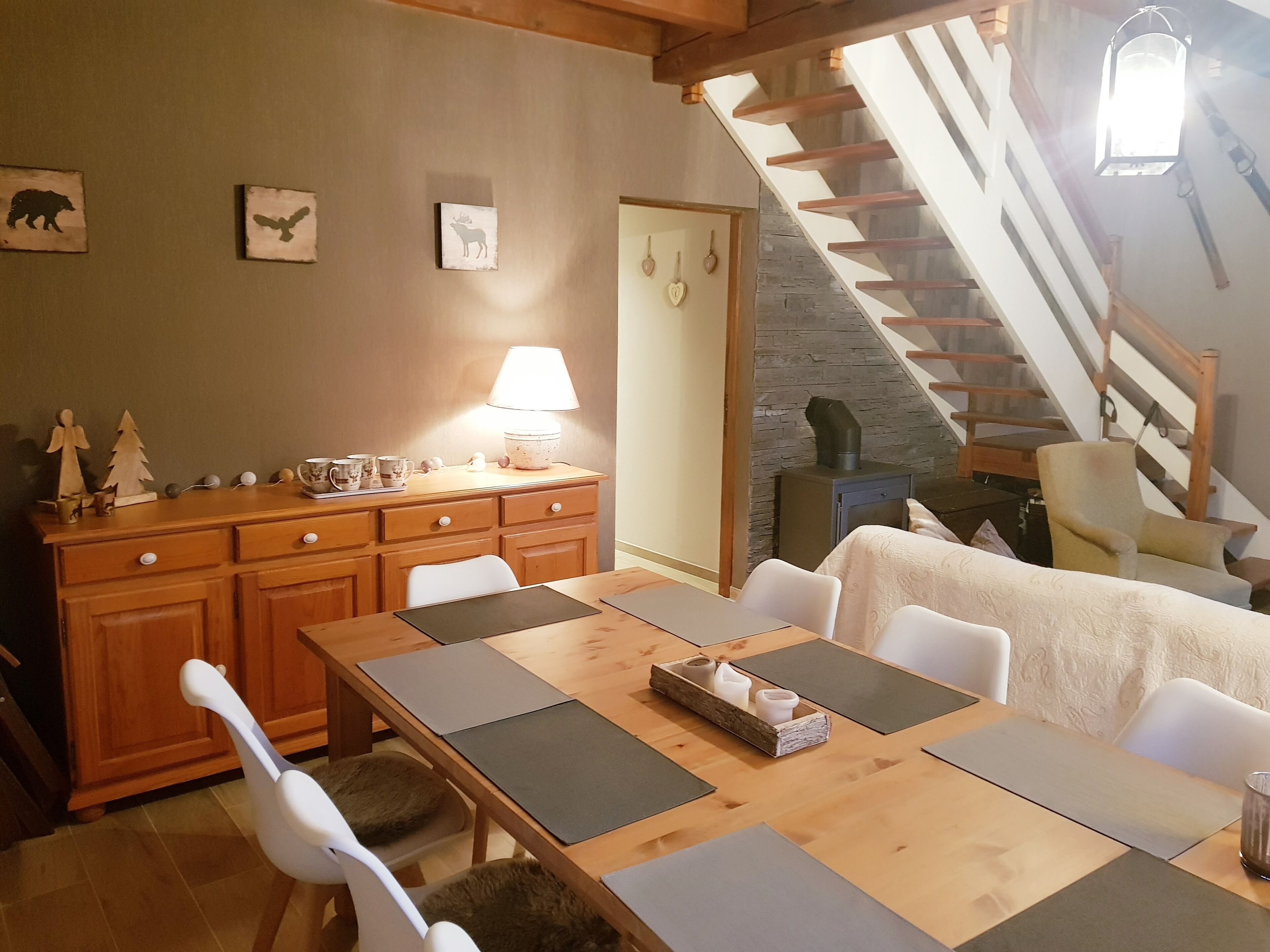 Un coin repas chaleureux avec une table en bois dressée pour quatre personnes, une lampe et un escalier menant à l’étage supérieur.