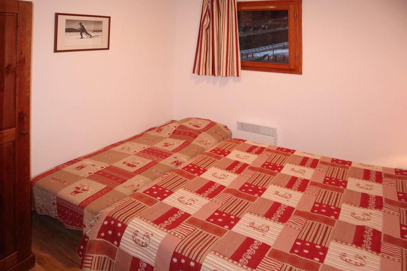 A single bed with a patterned quilt, a wooden door, a framed picture on the wall, and a window with curtains.