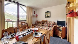 Una stanza accogliente con un tavolo da pranzo apparecchiato per due persone, un divano, una TV e vista sulle montagne.