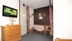 Et værelse med et trækommode, et fjernsyn ophængt på væggen, et lille bord med en stol og en seng med et mønstret sengetæppe.