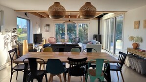 A modern dining area with a wooden table and chairs, large windows, and a view of greenery.