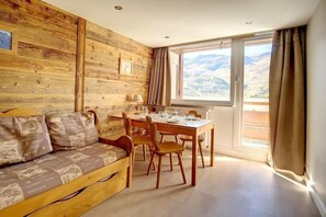 A cozy room with a wooden wall, a sofa with patterned cushions, a dining table with chairs, and a large window offering a mountain view. - 150m from resort center, 2nd floor, tv, 36m², Les Menuires (Les Menuires)