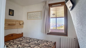 A bedroom with a wooden headboard, a patterned bedspread, a window with a view of mountains, a radiator, and a framed picture on the wall. - 250m from resort center, 8th floor, view mountain, balcony, tv, 25m², Les Menuires (Les Menuires)
