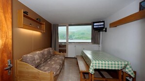 Ein kleines Zimmer mit einem Holzbett, einer Bank mit kariertem Tischläufer, einem Fernseher an der Wand und einem Fenster mit Blick auf Grünflächen.