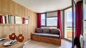 A room with a wooden wall, a built-in bench with cushions, a dining table with cutlery, and a large window with red curtains. - 2nd floor, balcony, tv, ski locker, 24m², Les Menuires (Les Menuires)