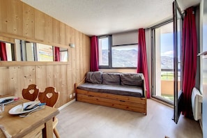 Een kamer met een houten wand, een ingebouwde bank met kussens, een eettafel met bestek, en een groot raam met rode gordijnen.
