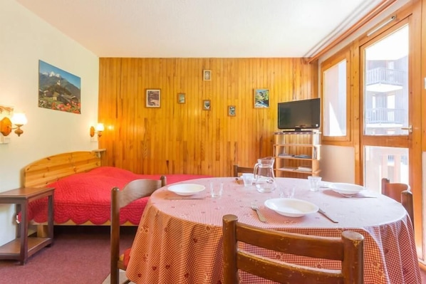 Una habitación con pared de madera, una cama con manta roja, una mesa de comedor dispuesta con platos y vasos, y un televisor sobre un mueble.