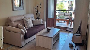 Sala de estar com sofá, mesa de centro e uma mesinha de cabeceira com abajur. Há uma varanda com vista para edifícios e áreas verdes.