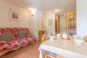 A cozy living room with a red and white patterned sofa, a dining table with chairs, and a bookshelf.