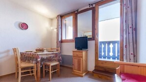 Une pièce chaleureuse avec une table en bois, des chaises, un téléviseur posé sur un meuble et un balcon doté d’une balustrade.