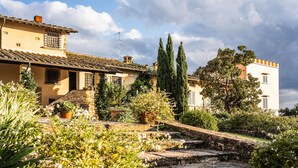 Ein Gebäude im traditionellen mediterranen Stil mit einer Steintreppe, die zum Eingang führt, umgeben von üppiger Vegetation und blühenden Pflanzen.