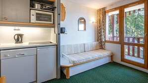 Une petite cuisine équipée d’un évier, d’une cuisinière et d’un petit réfrigérateur. On y trouve un banc avec un coussin et une fenêtre avec des rideaux.