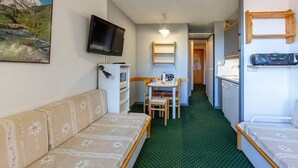 Une chambre d’hôtel compacte comprenant un canapé, une petite table à manger, une télévision, un réfrigérateur et une étagère en bois.