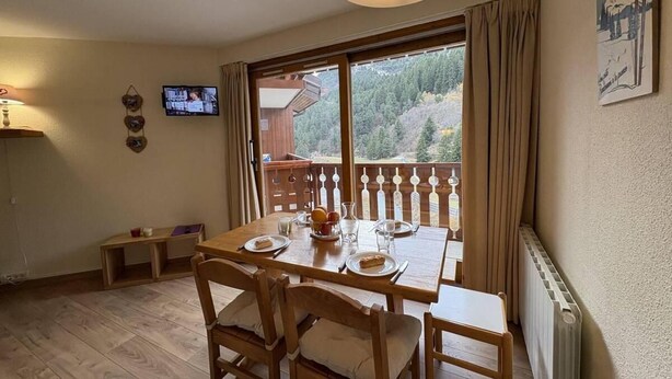 A dining area with a table set for two, a television on the wall, and a view of a forested area through large windows. - 300m from resort center, 3rd floor, view mountain, balcony, 27m², Méribel-Mottaret (Méribel - Les Allues)