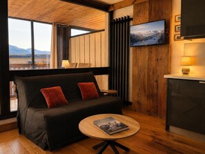 A modern room with a black sofa, a round coffee table with a book, a TV on the wall, and a view of mountains through large windows.