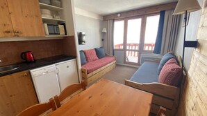 Cocina pequeña con armarios de madera y una mesa de comedor. Hay un sofá con cojines y una ventana con cortinas.