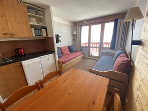 A compact kitchen with wooden cabinets and a dining table. There is a sofa with cushions and a window with curtains. - Ski-in/ski-out & foot of the slopes, 5th floor, view ski slopes, balcony, tv, ski locker, 27m² (La Plagne)