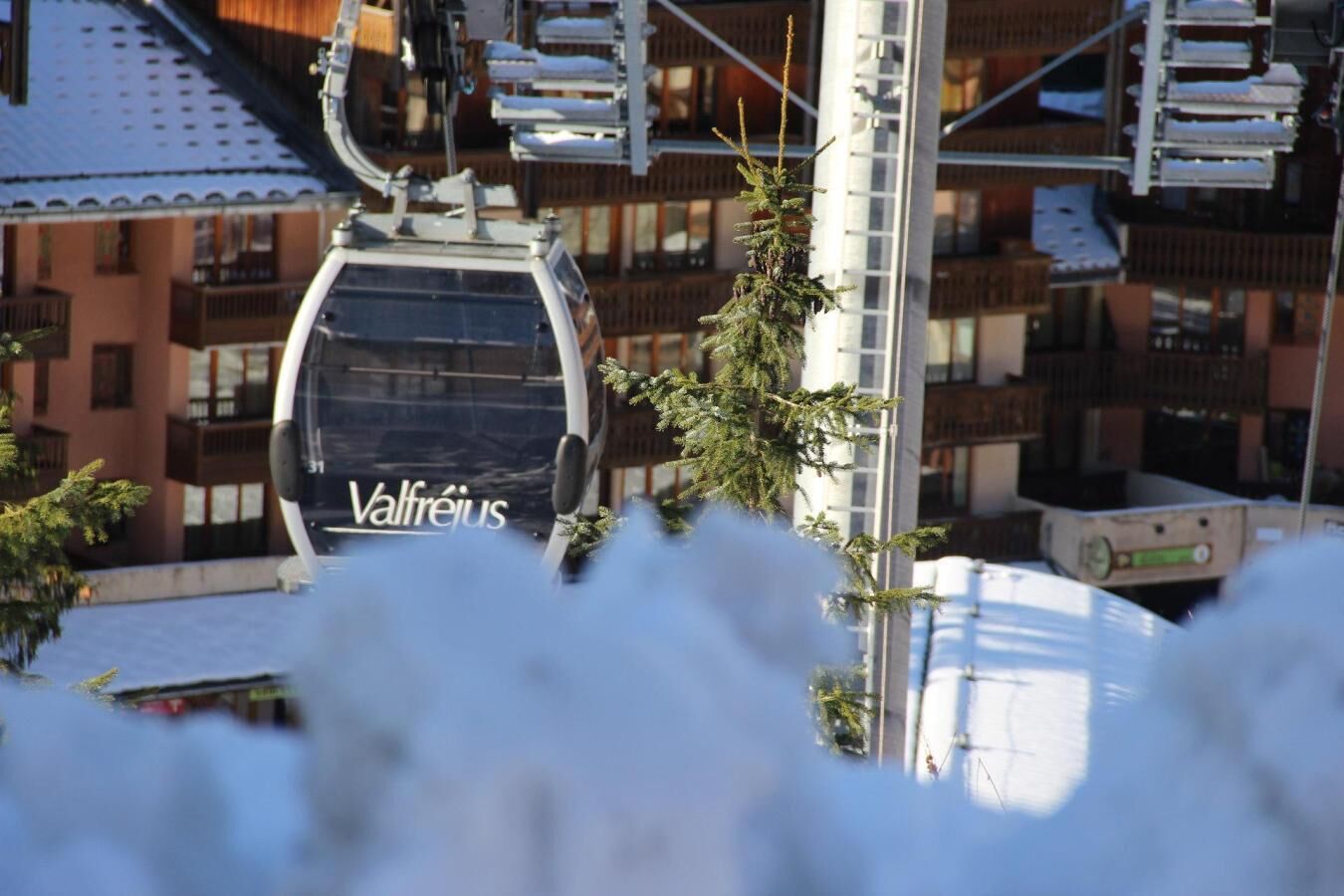 Eine Seilbahn mit dem Schriftzug 'Valfréjus', ein Gebäude mit Balkonen und ein Baum im Vordergrund.